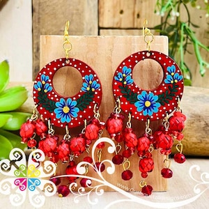 May include: Red, round earrings with a floral design. The earrings feature a central hole, blue flowers, white polka dots, and red beaded accents. Gold-colored hooks and dangling red beads complete the design.