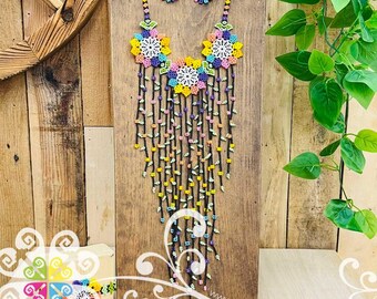 May include: A colorful beaded necklace and matching earrings set. The necklace features large floral designs in yellow, pink, and blue, with long beaded strands. The earrings mirror the floral design. The jewelry is displayed on a wooden stand.