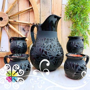 May include: A black ceramic pitcher and four matching mugs. The pitcher has a wide base and a handle, with a decorative floral pattern. The mugs also feature the same floral design. The set is displayed on a white surface.