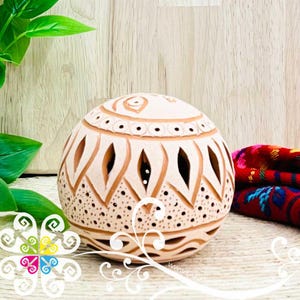May include: A spherical, off-white ceramic candle holder with intricate cut-out designs. The decorative piece features geometric patterns and small circular perforations. The candle holder is placed on a textured surface, with a glimpse of colorful textiles in the background.