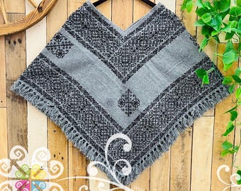 May include: A grey poncho featuring intricate black embroidered floral and geometric patterns along the V-neckline and hem. The garment has a V-shaped silhouette with a fringed edge, showcasing detailed needlework across the front surface.