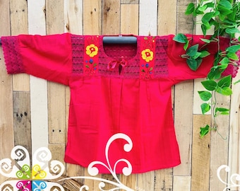 May include: A vibrant fuchsia-colored blouse with embroidered floral details. The top features a square neckline with a small bow, short sleeves with lace trim, and yellow and blue floral embroidery. The blouse is displayed against a wooden background.