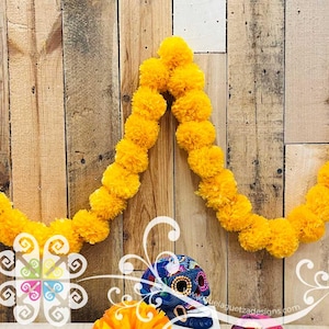 May include: A vibrant yellow marigold flower garland draped in a V-shape, featuring dense, ruffled pom-pom blossoms. Below the garland sits a decorative blue skull with intricate multi-colored floral patterns and the text GuelaguetzaDesigns.