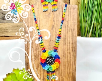 May include: A colorful beaded necklace and earrings set with a large flower pendant. The necklace is made of small, colorful beads and features a large, multi-colored flower pendant with a black center. The earrings are also made of small, colorful beads and feature a smaller flower design.