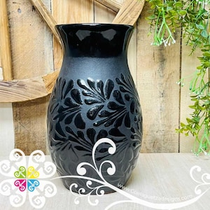 May include: A black ceramic vase with a matte finish and embossed floral design. The vase has a wide base that tapers to a narrower neck and a flared rim. The floral pattern is repeated around the body of the vase.