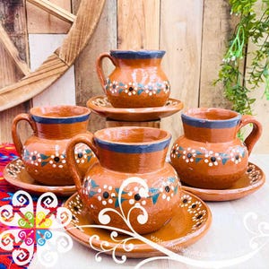 May include: Set of four terracotta mugs with matching saucers. Each mug has a blue rim and handle, with a floral design in white, blue, and black. The saucers have a matching design. The mugs and saucers are arranged on a wooden surface.
