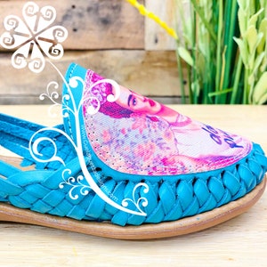 Pode incluir: Close-up de uma sand&aacute;lia de couro azul turquesa com um design tecido. A sand&aacute;lia apresenta uma imagem colorida de Frida Kahlo na ponta. O texto "www.CuelaguetzaDesigns.com" est&aacute; impresso na lateral da sand&aacute;lia.