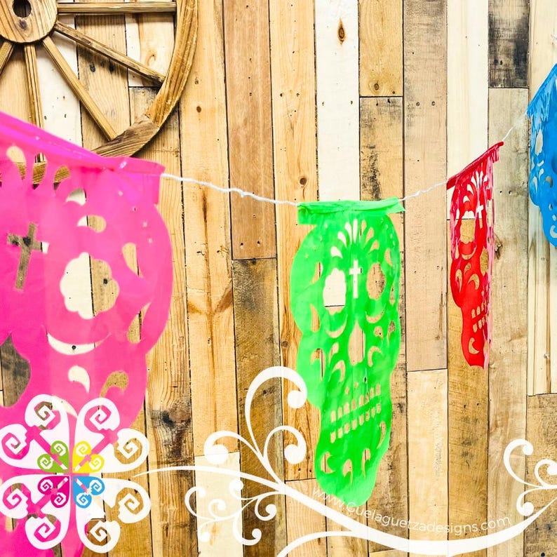 K&ouml;nnte beinhalten: Ein farbenfrohes Papierbanner mit ausgeschnittenen Zucker Sch&auml;del-Designs in Pink, Gr&uuml;n und Rot. Das Banner ist an einer wei&szlig;en Schnur befestigt und h&auml;ngt vor einem Holz Hintergrund.