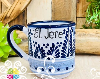 Op de afbeelding: Witte keramische mok met blauwe bloemmotieven en de tekst "El Jefe". De mok heeft een blauw handvat en rand. Het ontwerp omvat gestileerde bladeren en stippen. De mok staat op een houten oppervlak.