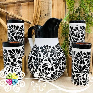 May include: A black and white ceramic pitcher and matching glasses set. The pitcher and glasses feature a floral design. The pitcher has a black top and handle, with a white band around the middle. The glasses have a black rim and a floral pattern.