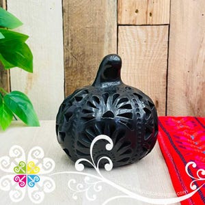May include: A black ceramic decorative gourd-shaped object with intricate cut-out floral designs. The gourd has a small, rounded top and sits on a white surface. A red and pink textile is in the background. The website address www.Guelaguetzadesigns.com is visible.