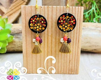Pode incluir: Brincos artesanais com pingentes circulares pretos adornados com desenhos florais vermelhos, laranjas e amarelos. Cada brinco tem uma borla e detalhes de contas em tons de rosa, roxo e branco, suspensos por ganchos dourados.