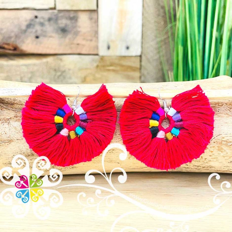 May include: A pair of vibrant red tassel earrings. Each earring features a fan-shaped design with a central ring adorned with colorful beads. The earrings hang from silver-colored hooks, offering a bold and eye-catching accessory.