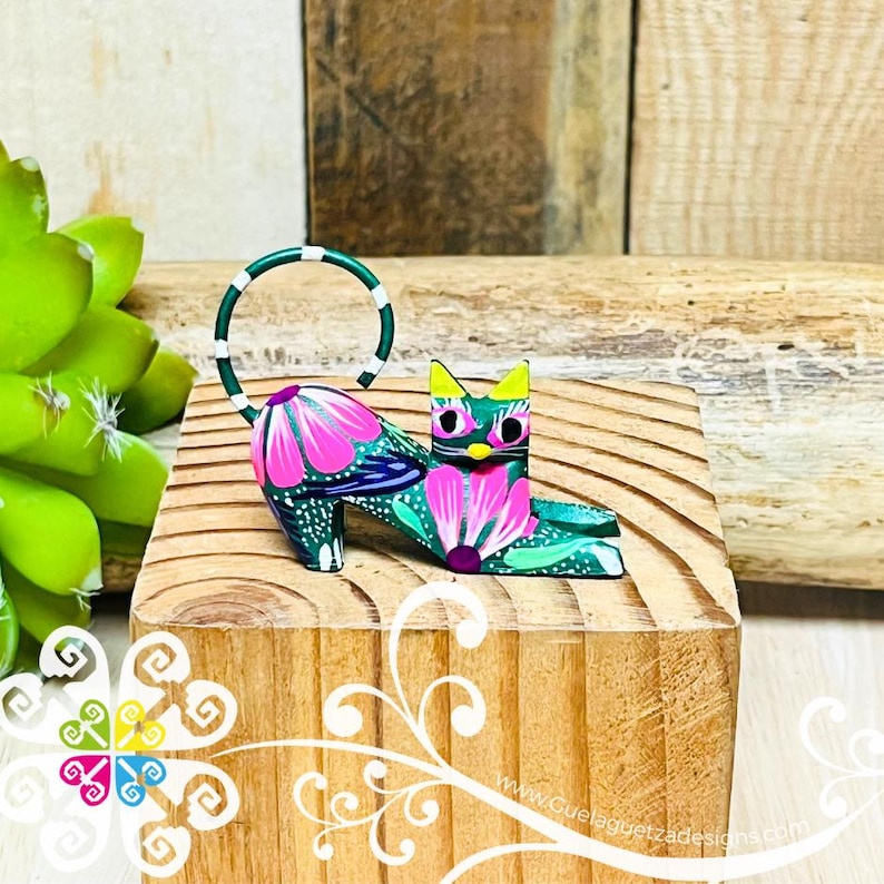 May include: A green, pink, and white hand-painted wooden cat figurine. The cat is lying down with its tail curled up. The cat has a yellow nose and green eyes.