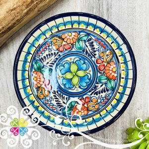 May include: A hand-painted ceramic plate with a vibrant floral design. The plate features a blue rim and a central design of orange, red, and blue flowers with green leaves. The background is cream-colored with a repeating pattern. The website address "Quelaguerzadesigns.com" is visible.