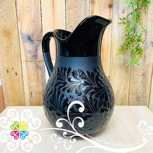 May include: A black ceramic pitcher with a glossy finish. The pitcher features a matte band around the middle and a detailed floral pattern. The handle is curved, and the spout is wide. The pitcher is displayed on a wooden surface.
