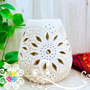 May include: Cream-colored ceramic vase with a floral design. The vase has cut-out patterns and small holes, creating a decorative effect. The design is centered on the vase, with a series of petal-like shapes. The vase is on a woven surface.