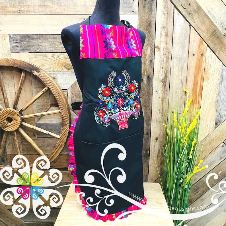 May include: Black apron with a pink and multi-colored embroidered floral design. The apron has a ruffle trim and a pocket. The text "www.crazedesigns.com" is visible in the bottom right corner.