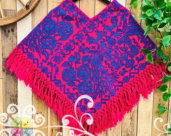 May include: A bright pink poncho featuring a dense, intricate blue woven pattern of floral and bird motifs. The garment has a V-neckline and is finished with long, matching pink fringe along the bottom edge, creating a textured appearance.