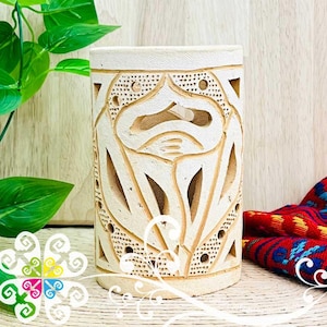 May include: A cream-colored, cylindrical candle holder with intricate carved floral and geometric patterns. The design includes cut-out shapes and small dotted details. The candle holder is displayed on a light-colored surface with a colorful textile in the background.