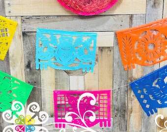 May include: A string of colorful paper banners with cut-out designs, including a flower, a jar, and a bird. The banners are strung on a white string and hanging against a wooden background. The text "www.GuadalupeZadesigns.com" is visible at the bottom of the image.