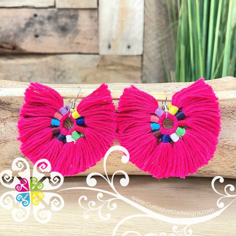 May include: A pair of vibrant fuchsia tassel earrings. Each earring features a fan-shaped design with a central ring adorned with colorful beads. The earrings are suspended from silver-colored hooks.