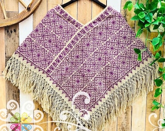 May include: A beige poncho featuring a repeating deep purple geometric floral embroidery pattern across the fabric. The garment has a V-shaped neckline and is finished with long, thick beige fringe along the bottom hem, hanging from a black hanger.
