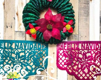 May include: Two teal and magenta paper banners with the text "Feliz Navidad" in white. The banners are decorated with cut-out designs of poinsettias and a grid pattern. A green wreath with red poinsettias is in the background.
