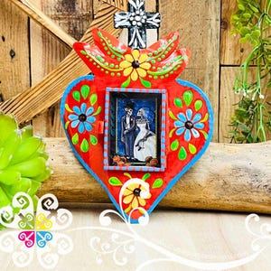 May include: A vibrant, heart-shaped Day of the Dead wall hanging. The red metal art piece features a central framed image of a skeleton couple, surrounded by painted flowers and a cross. The piece is approximately 8 inches wide.