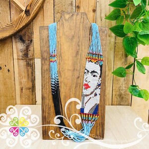 K&ouml;nnte beinhalten: Eine Perlenkette mit einem Portr&auml;t von Frida Kahlo. Die Kette hat einen hellblauen Perlenstrang, der in schwarze Perlen &uuml;bergeht. Das Portr&auml;t ist wei&szlig; mit roten Lippen und einem blau-gelben Kopfschmuck. Die Kette wird auf einem Holzst&auml;nder pr&auml;sentiert.
