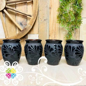 May include: Four black ceramic vases with a matte finish and glossy black floral designs. The vases have a rounded shape and a dark rim. The background includes a wooden wheel and wooden planks.