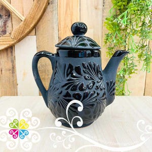 May include: Black ceramic teapot with a lid and handle. The teapot features a raised floral design and a glossy finish. The spout and handle are curved. The teapot is sitting on a white surface with a wooden background.
