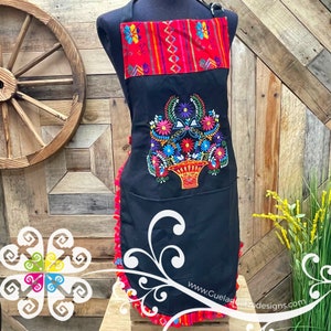 May include: Black apron with a red and orange striped waistband and a colorful embroidered floral design. The apron has a ruffled bottom and a pocket on the front. The text "www.GuelaguetzaDesigns.com" is printed on the bottom of the apron.