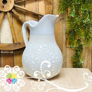 May include: White ceramic pitcher with a white floral design. The pitcher has a wide mouth and a handle.
