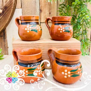May include: Four terracotta mugs with handles, featuring a rustic design. Each mug is a warm orange-brown color, adorned with white dots and green leaf-like patterns. The mugs are displayed on a wooden surface, showcasing their traditional craftsmanship.