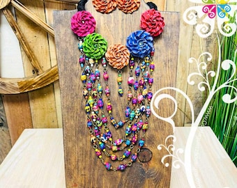 May include: A vibrant necklace and earring set displayed on a wooden stand. The necklace features layered strands of colorful beads and is adorned with large, fabric flower accents in shades of pink, green, orange, and blue. The matching earrings echo the flower design.