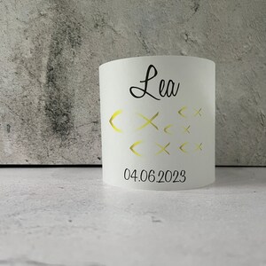 May include: White cylindrical candle holder with the name "Lea" and the date "04.06.2023" printed in black. The candle holder has a design of gold fish cutouts.