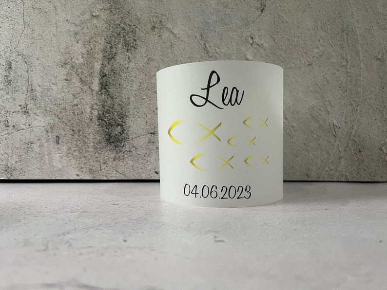 May include: White cylindrical candle holder with the name "Lea" and the date "04.06.2023" printed in black. The candle holder has a design of gold fish cutouts.
