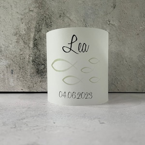 May include: White cylindrical candle holder with a cutout design of fish and the name "Lea" and the date "04.06.2023" printed in black.