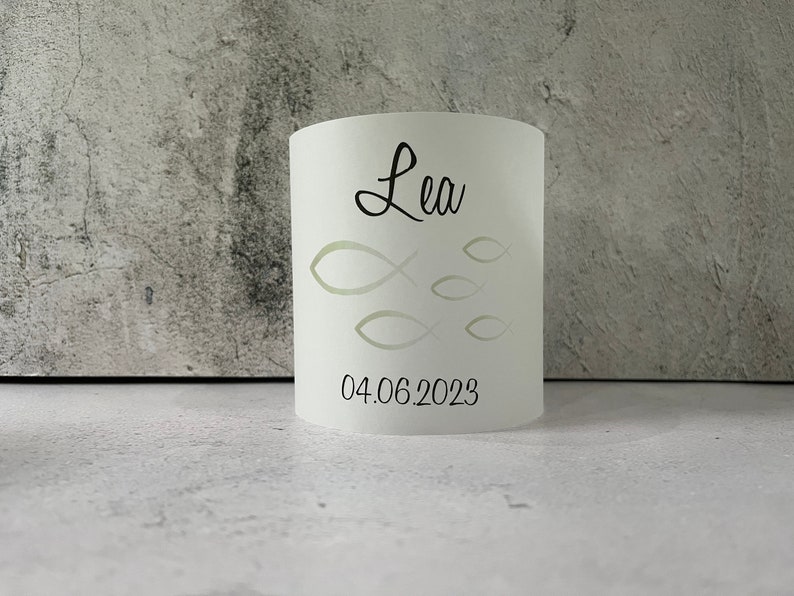 May include: White cylindrical candle holder with a cutout design of fish and the name "Lea" and the date "04.06.2023" printed in black.