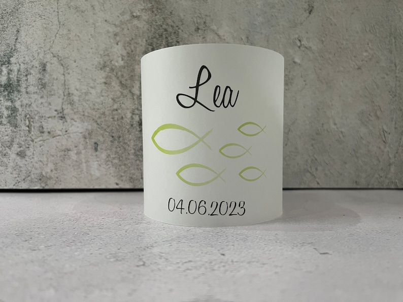 May include: White cylindrical candle holder with a green fish design and the name "Lea" and the date "04.06.2023" printed on it.