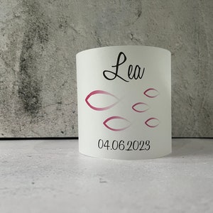May include: White cylindrical candle holder with a pink and white fish design and the name "Lea" and the date "04.06.2023" printed on it.