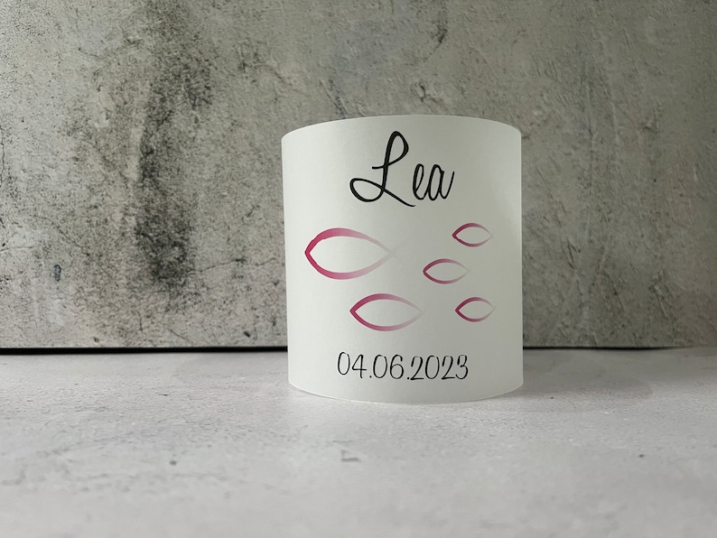 May include: White cylindrical candle holder with a pink and white fish design and the name "Lea" and the date "04.06.2023" printed on it.