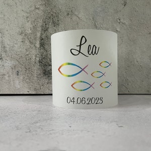 May include: White cylindrical candle holder with a rainbow-colored fish design and the name "Lea" and the date "04.06.2023" printed on it.