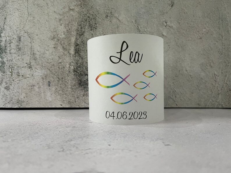 May include: White cylindrical candle holder with a rainbow-colored fish design and the name "Lea" and the date "04.06.2023" printed on it.