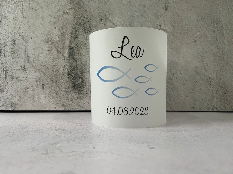 May include: White cylindrical candle holder with a blue and white fish design and the name "Lea" and the date "04.06.2023" printed on it.