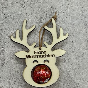Puede incluir: Adorno navide&ntilde;o de madera con forma de reno con la inscripci&oacute;n alemana "Frohe Weihnachten" y un bomb&oacute;n Lindor. El adorno tiene un acabado de madera natural y una cuerda marr&oacute;n para colgar. Los cuernos y la cara del reno est&aacute;n recortados.
