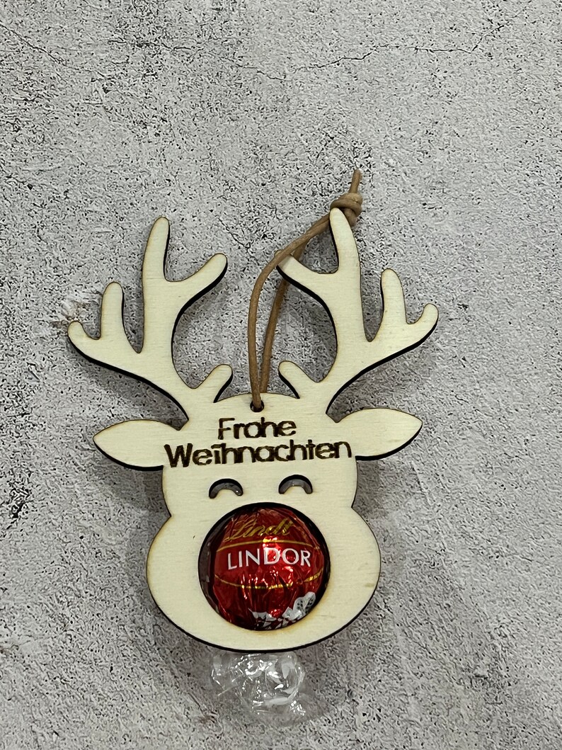 Puede incluir: Adorno navide&ntilde;o de madera con forma de reno con la inscripci&oacute;n alemana "Frohe Weihnachten" y un bomb&oacute;n Lindor. El adorno tiene un acabado de madera natural y una cuerda marr&oacute;n para colgar. Los cuernos y la cara del reno est&aacute;n recortados.