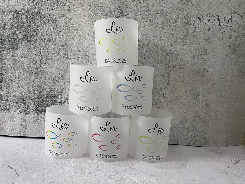 May include: Six white cylindrical candle holders with a design of a school of fish and the name "Lea" and the date "04.06.2023" printed on each.