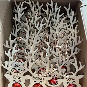 Puede incluir: Adornos navide&ntilde;os de renos de madera, cada uno con un nombre grabado en las astas. Las caras de los renos contienen bombones Lindt Lindor. Los adornos est&aacute;n en una caja de cart&oacute;n.